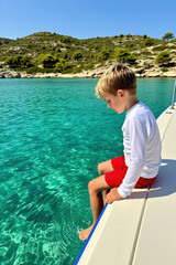 Fototapeta premium Boy sits on boat edge dipping feet in clear turquoise sea. Sunny day, islands with green bushes, blue sky. Child enjoys summer boat trip vacation.