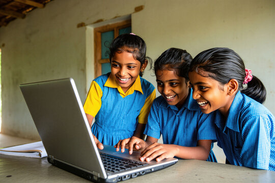 Three smiling Indian schoolgirls in blue uniforms use laptop computer together in classroom. Students learn digital skills on portable pc during lesson at rural government school. Children enjoy