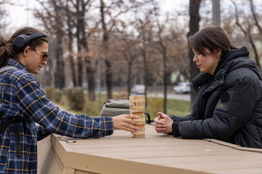 A happy female student laughing and playing a board game with her classmate in a city park. Genuine emotions of student life and friendship.