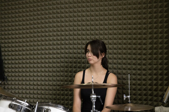 Young woman music college student sitting at a white drum kit, looking aside in a rehearsal room with pyramid foam walls.