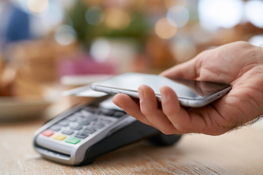 Hand holds smartphone above payment terminal. Blurred background suggests cafe or shop setting. Focused on contactless transaction moment. Implies modern, convenient, digital payment method