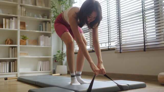 Woman doing deep split stretch with strap on pilates mat, low angle close-up flexibility exercise, home fitness studio with bookshelf and dumbbells, advanced stretching wellness scene