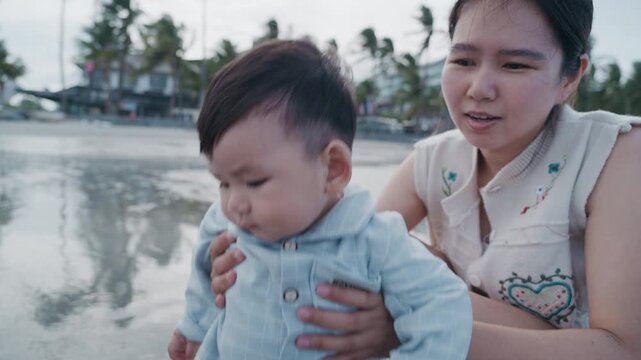 Happy Asian mother holding her cute baby boy on the tropical beach.