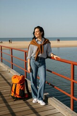Fototapeta premium Woman with orange backpack stands on wooden pier. She smiles, enjoying sunny coastal weather. Casual outfit suits seaside travel and beach walks. Summer vacation vibes.