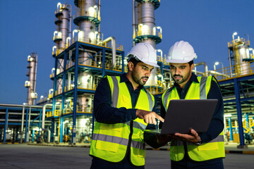 Fototapeta premium Two engineers wear hard hats and safety vests. They look at laptop screen with refinery plant at night. People work together, discuss project.