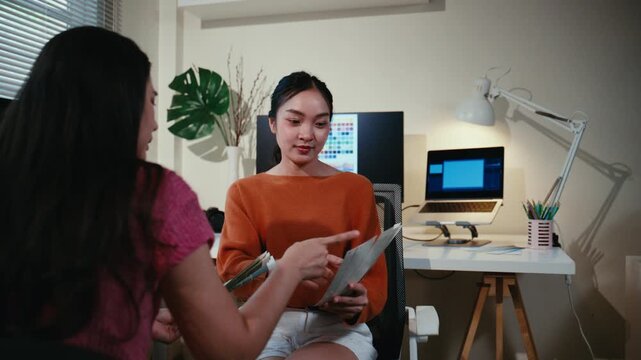 Two young Asian female graphic designers discussing a project and choosing colors from a palette sample in a modern office at night