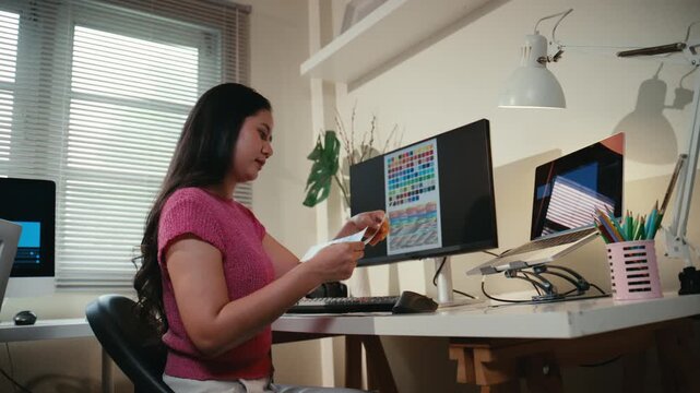Young Asian woman working at her desk. Professional graphic designer or video editor comparing colors on a swatch with a computer monitor