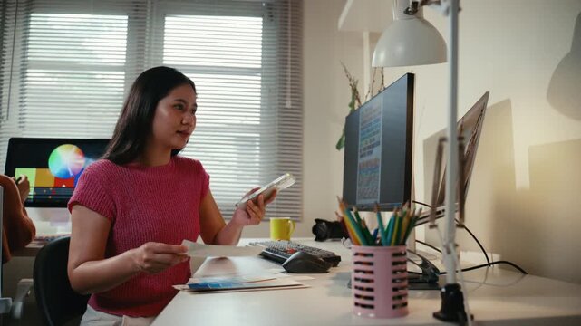 Young Asian woman working at her desk, checking color swatches and using her phone for reference. A professional creative in her office