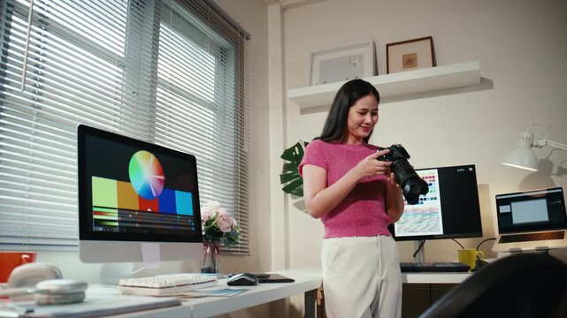 Young Asian female photographer checking her camera while working on a desktop computer with multiple displays for photo editing