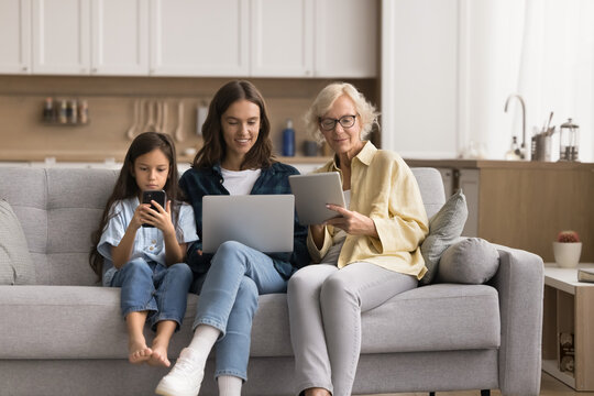 Absorbed senior 60s granny, her grown-up 30s daughter and pre-teen 10s granddaughter using diverse wireless devices seated on couch in living room. Modern technology usage addiction, bad habit, hobby