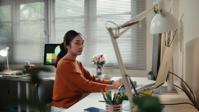 Concentrated young Asian woman typing on a keyboard, working on a computer and laptop at her creative home office desk during the day