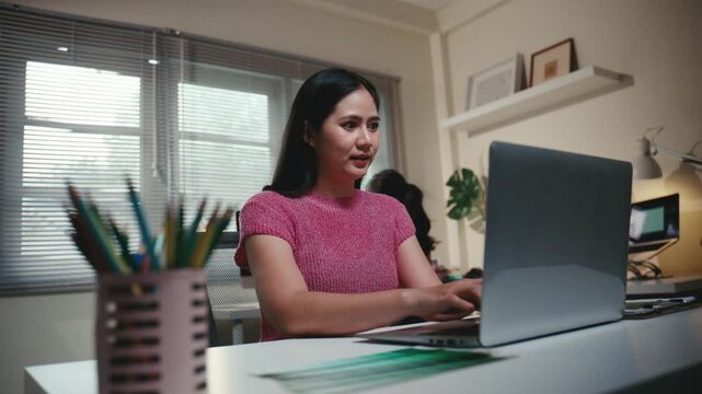 Young Asian woman working late at her home office, typing on a laptop computer. Focused female designer creating a project at night
