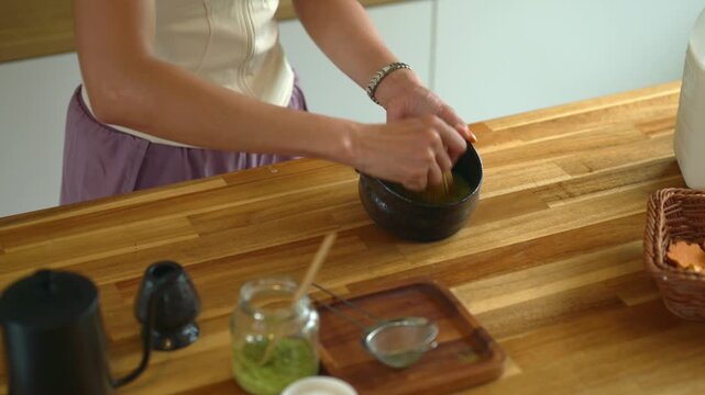 Woman Whisking Matcha Green Tea in Traditional Black Bowl on Wooden Kitchen Counter