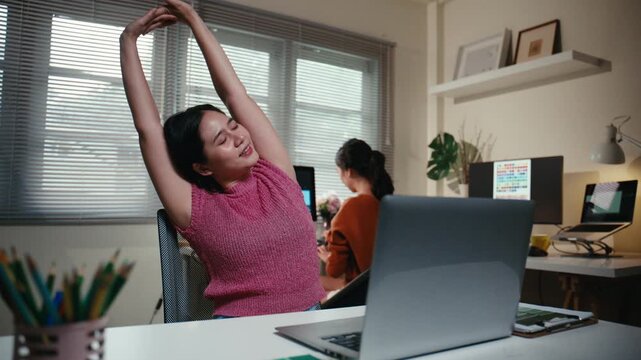 Young Asian woman feeling tired and taking a break, stretching her arms and body to relieve muscle tension after long hours at her desk