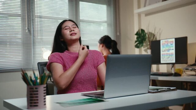 Young Asian woman feeling tired and stressed while working late at her desk, experiencing neck pain and office syndrome from overwork