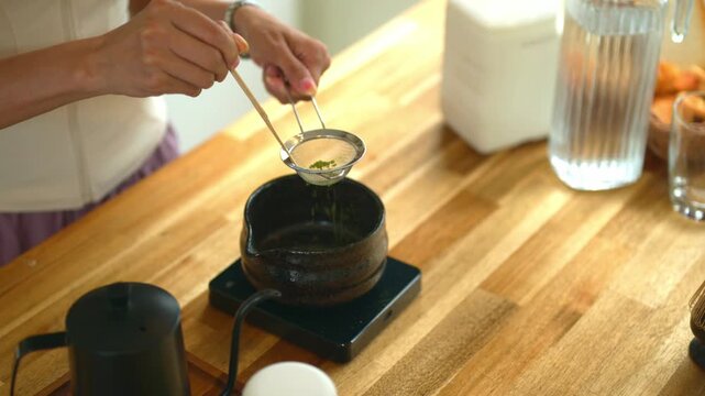 Closeup Woman uses bamboo scoop to sift vibrant green matcha powder through a fine mesh strainer into a dark ceramic