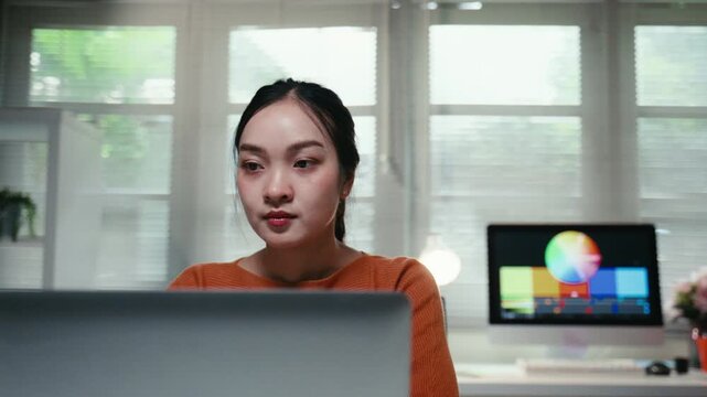 Young Asian woman working from home office as a video editor or colorist. A dedicated creator using a laptop and an external monitor for her project