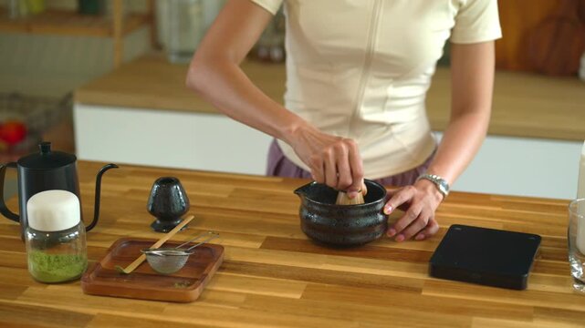 Woman Preparing Matcha Tea with Bamboo Whisk and Ceramic Bowl on Wooden Kitchen Counter