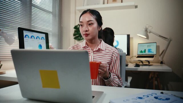 Young Asian businesswoman holding a coffee mug while analyzing financial data on her laptop and tablet, working late in the office