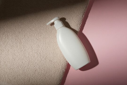White cosmetic bottle with dispenser pump rests on soft textile towel over pastel pink surface. Minimalist skincare packaging layout shows empty plastic container for liquid soap lotion hair shampoo.