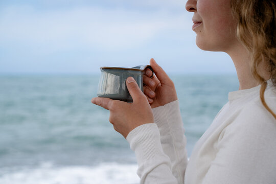 Cropped view of a Caucasian female holding a hot beverage on a cloudy day near the ocean. Hygge, relaxation, and coastal vacation concepts.
