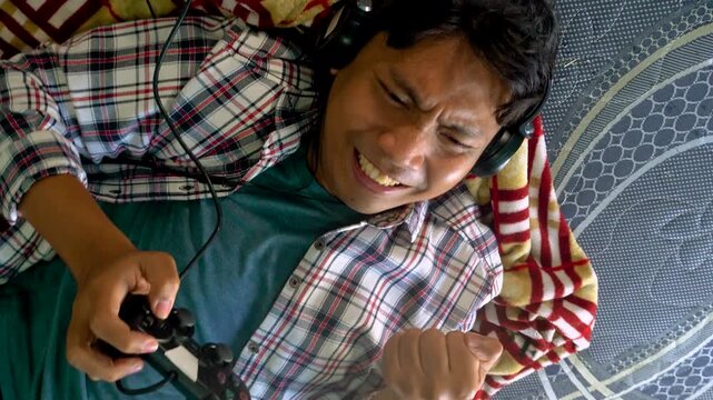 Young Man Playing Video Games With Headphones Celebrating Success While Lying On Patterned Bedding