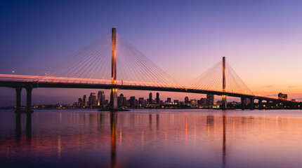 Fototapeta premium Wide angle cable-stayed bridge with tall pylons reflecting on still river under vivid purple pink sunset sky with distant city skyline, representing iconic urban landmark and travel destination