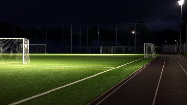 Cinematic night view of an empty football stadium with illuminated soccer goals, vibrant artificial turf, and a running track under powerful floodlights. The quiet sports arena creates a dramatic