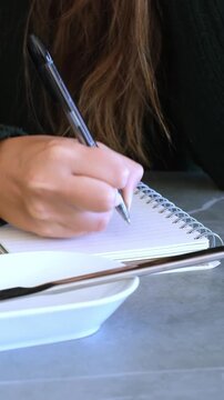 A close-up shot captures a person's hand diligently writing notes in a spiral notebook with a pen, set on a grey table alongside a white bowl. Ideal for themes of study, work, planning, and creative t