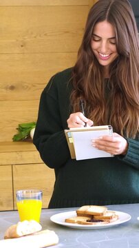 A smiling young woman writes in her notebook in a modern kitchen, planning her day. Toast and orange juice nearby, she embraces a mindful morning routine, reflecting domestic bliss and healthy living.