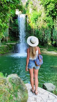 Back view of woman traveler with straw hat and backpack looking at tropical waterfall and emerald pool, Spain, Frias