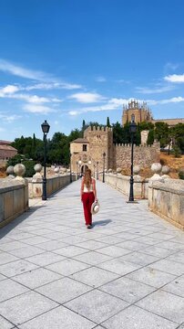 Back view of woman tourist in red pants walking on medieval stone bridge towards Toledo fortress in Spain