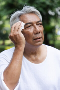 Tired and uncomfortable old asian senior man wiping his face with refreshing cold wet towel, trying to relieve from heavy sweating and outdoor summer heat wave