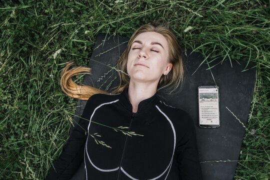 Woman lies on mat in grass with eyes closed next to smartphone. Somatic yoga practice, nervous system regulation, trauma informed yoga, mindbody connection.