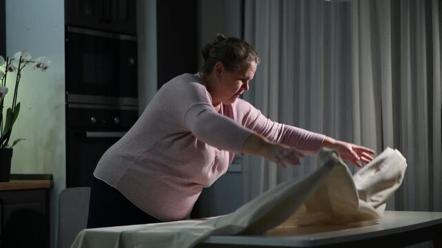 woman covering a dining table with a tablecloth in the evening. Cozy home preparation scene focused on household routine and table setting.