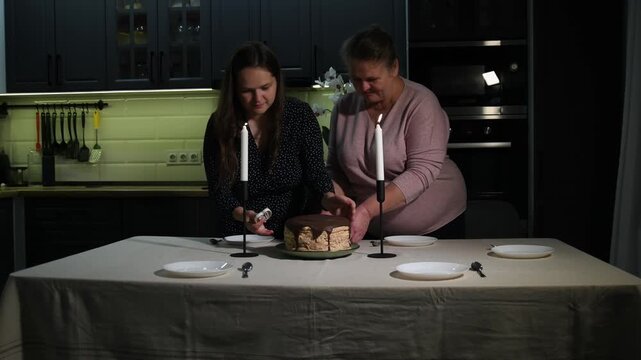 Two women place a birthday cake with number 60 candles on the table and add decorative candles, preparing a milestone celebration at home.