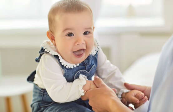 A joyful infant in denim overalls smiles while held, reaching for hands and interacting with a parent in a cozy home bathed in soft natural light. Baby giggles while holding parent's hands