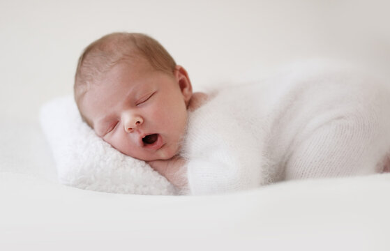 Newborn Sleeps Quietly On Fluffy White Pillow Wearing Cozy White Outfit And Hat With Hands Tucked Under Chin. Newborn Photo Shoot.