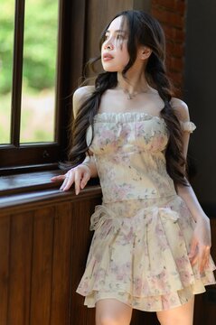 Young woman with long dark hair wearing a pastel floral dress by window indoors