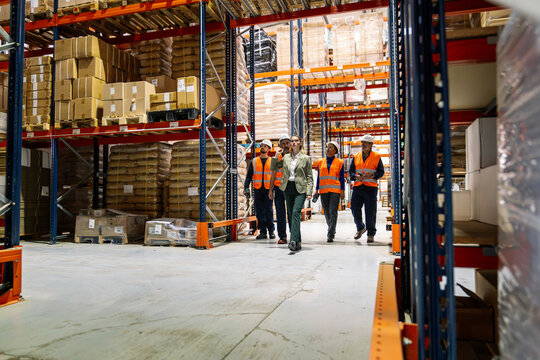 Business managers and workers walking and discussing operations in a busy warehouse aisle among storage shelves, showcasing teamwork and supply chain management