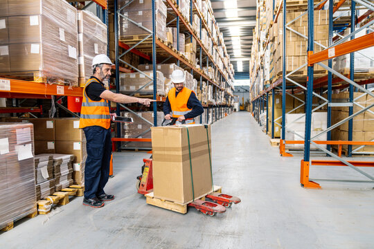 Two workers in safety vests and hard hats operating a pallet jack and checking inventory on a digital tablet in a large logistics warehouse