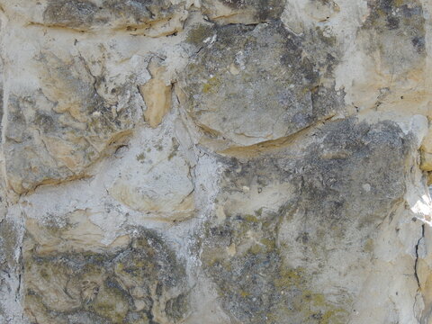An old stone wall made of uneven beige and gray cobblestones. The rough, textured surface reveals traces of light-colored cement mortar, a dark layer of weathering, and patches of moss.