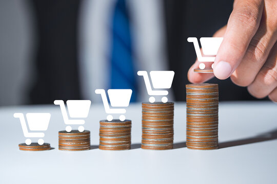 Businessman hand putting coin on growth chart stack with shopping cart icon, retail sales growth and e-commerce concept.