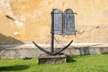 An ancient iron ship anchor on a stone pedestal against a weathered yellow colonial building wall with old wooden window shutters in Galle Fort, Sri Lanka. © Сергей Дудиков