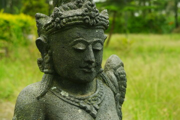 Fototapeta premium Ancient stone statue of a serene deity featuring intricate traditional carvings and mossy textures captured in a close up side profile shot set within a lush green tropical garden environment.