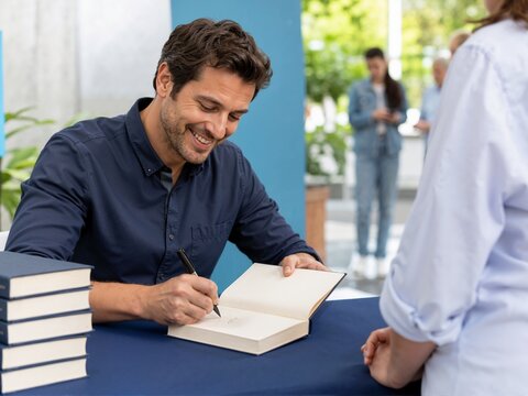 Smiling male author signing a book with a pen at a promotional event. Happy writer giving an autograph to a reader