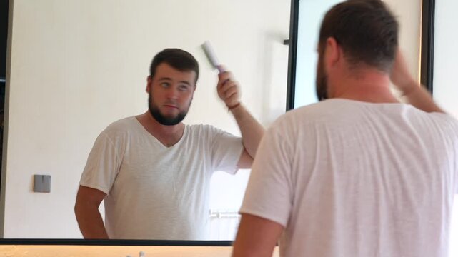 Young man standing in front of a mirror and combing his hair. Daily grooming routine at home, personal care, morning preparation, hygiene and self-care lifestyle.