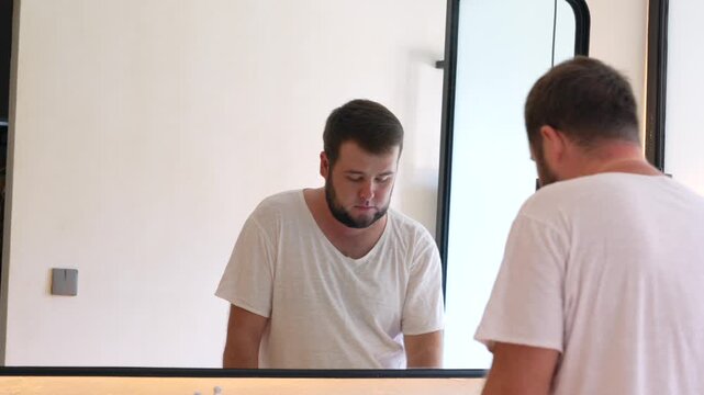 Young man carefully trimming his beard with an electric razor while looking in the mirror. Daily grooming routine, facial hair care, personal hygiene and self-care at home.