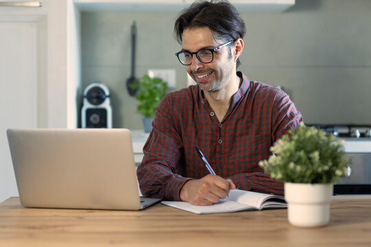 Happy man entrepreneur wearing eyeglasses looking at paper bill checking financial invoice or tax document making online payments working on laptop at home kitchen table.