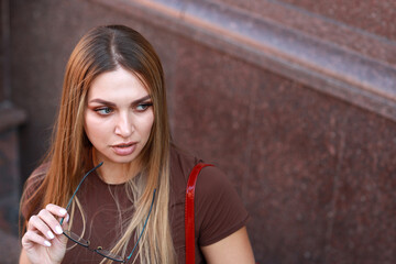 Fototapeta premium Close up portrait of a beautiful young woman with long light brown hair putting on eyeglasses while sitting near a granite wall in the city